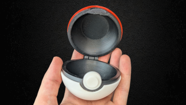 Hand holding a red and white 3D-printed Pokéball-style container with a black center band and circular button, against a black background.