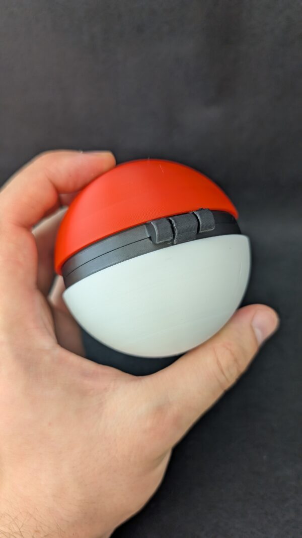 Hand holding a red and white 3D-printed Pokéball-style container with a black center band and circular button, against a black background.