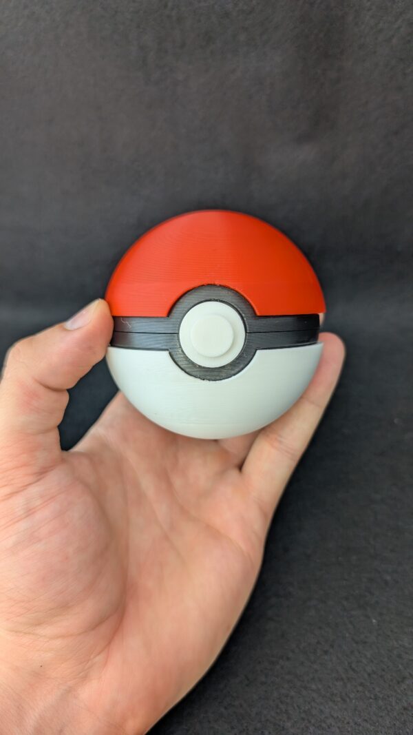 Hand holding a red and white 3D-printed Pokéball-style container with a black center band and circular button, against a black background.