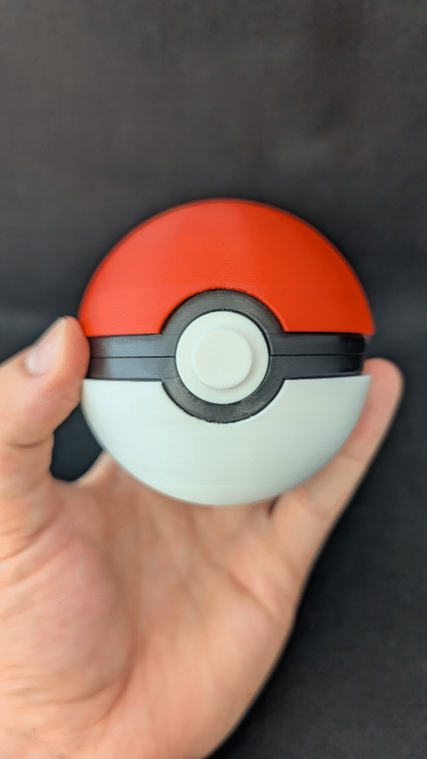 Hand holding a red and white 3D-printed Pokéball-style container with a black center band and circular button, against a black background.